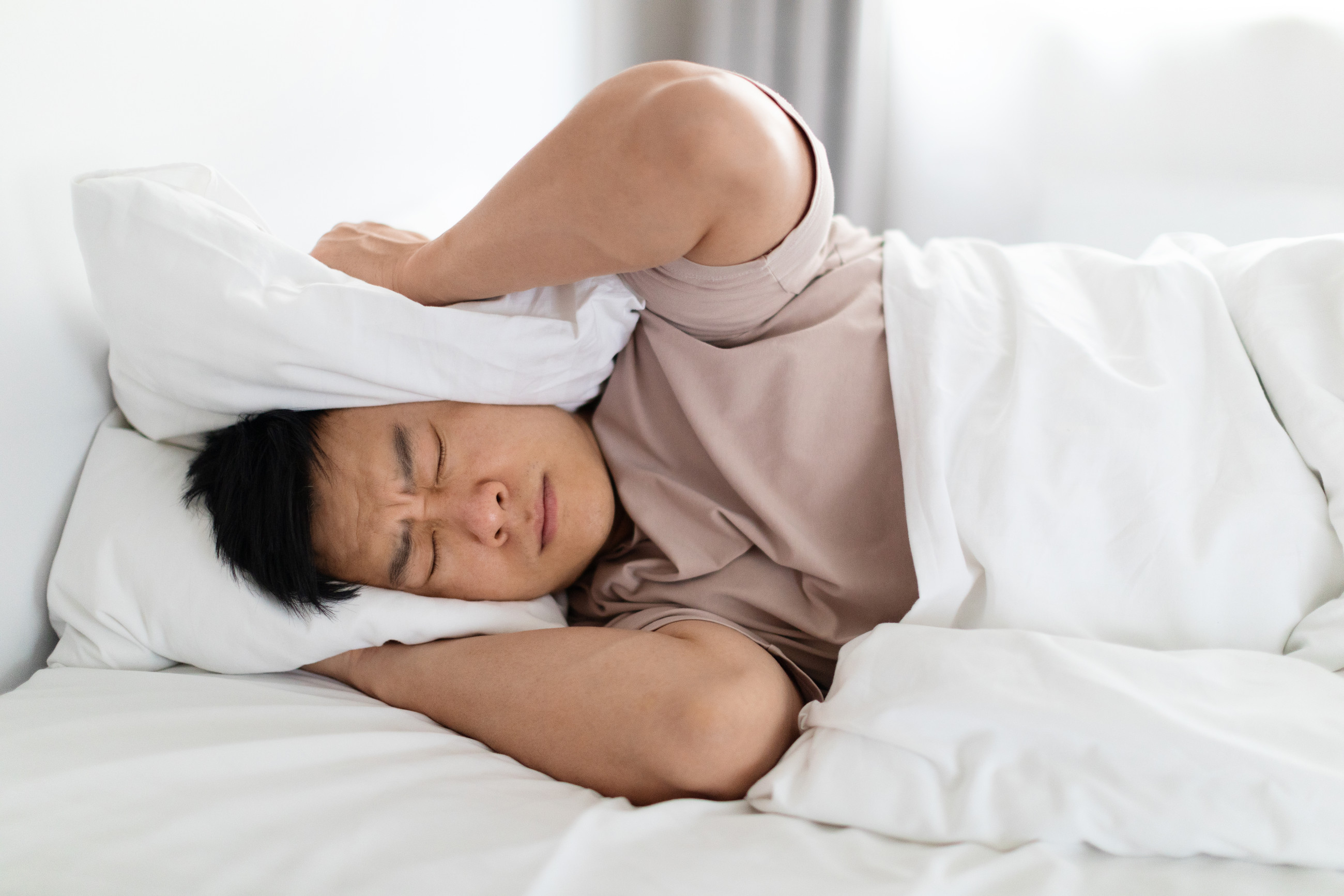 A man in bed holding a pillow over his ears from the noise outside